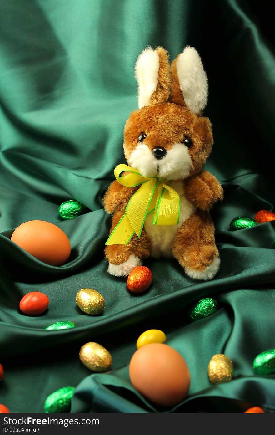 Easter theme - a fluffy bunny (rabbit) with a yellow ribbon sitting on a dark green satin fabric - textile, surrounded by colorful Easter eggs. Easter theme - a fluffy bunny (rabbit) with a yellow ribbon sitting on a dark green satin fabric - textile, surrounded by colorful Easter eggs.