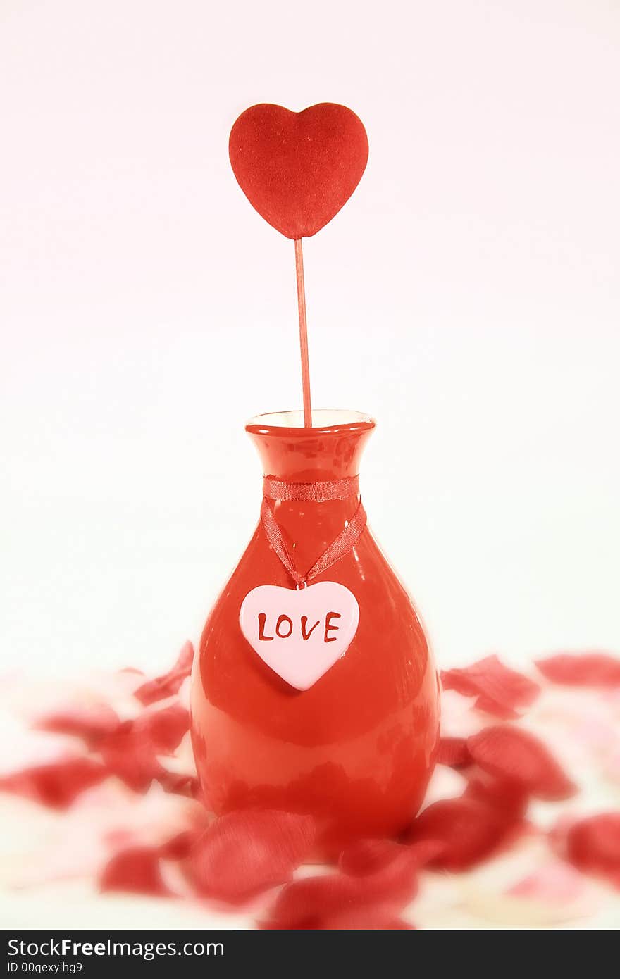 A red heart in a coordinating vase with love tag surrounded by rose petals with a gradated pale pink background. A red heart in a coordinating vase with love tag surrounded by rose petals with a gradated pale pink background.