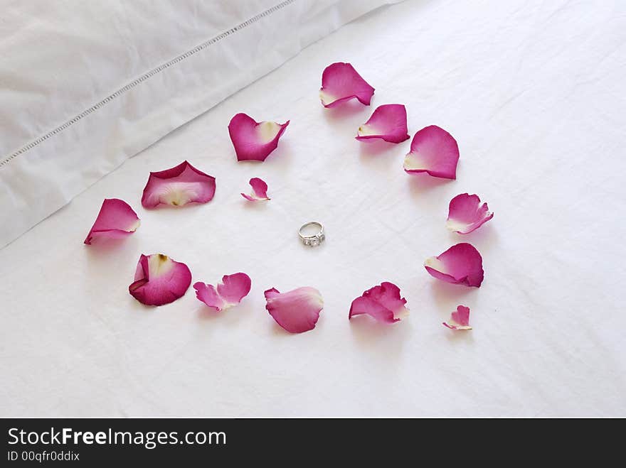 A pink heart made of rose petals on a crisp white bed enclose a diamond engagment ring. A pink heart made of rose petals on a crisp white bed enclose a diamond engagment ring