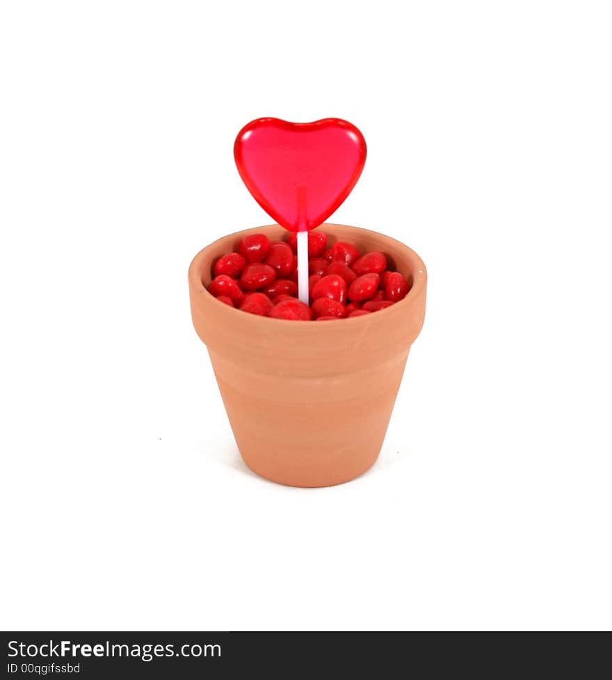 A red, heart shaped lollipop grows out of cinnamon heart soil in a terracotta flowerpot. Isolated on white background. A red, heart shaped lollipop grows out of cinnamon heart soil in a terracotta flowerpot. Isolated on white background.