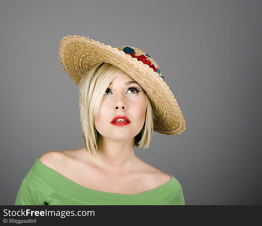 A blonde model in green blouse on a grey background with a flowered hat looking up. A blonde model in green blouse on a grey background with a flowered hat looking up