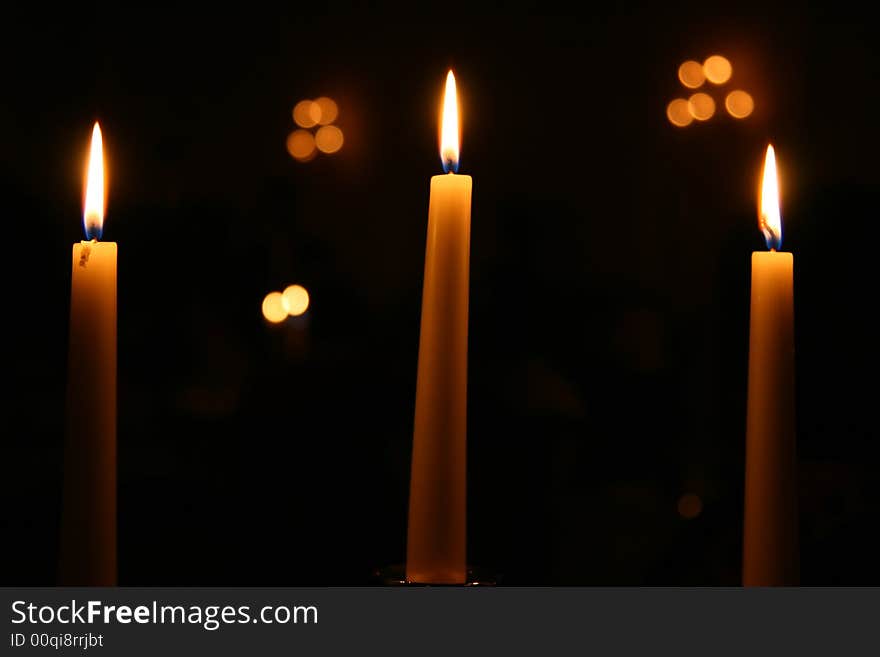 Three candles burning at night with other lights out of focus in the background