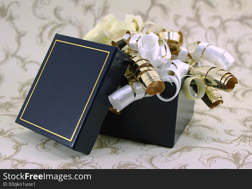 Curly gold, white and yellow ribbons spill out of a navy blue gift box. Curly gold, white and yellow ribbons spill out of a navy blue gift box.