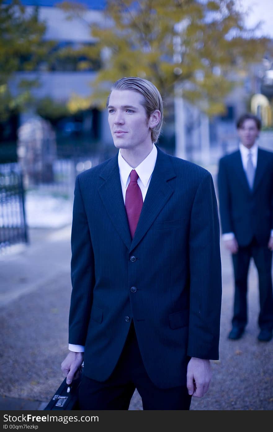 Two businessmen staggered one behind the other in full suits with one man holding briefcase. Two businessmen staggered one behind the other in full suits with one man holding briefcase