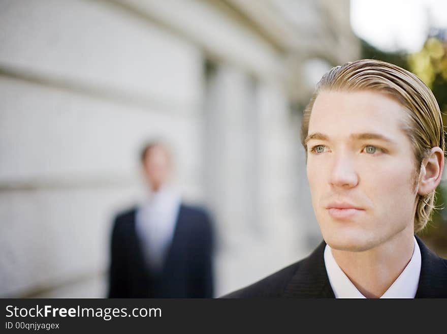 Two businessmen standing in front of building wearing suits looking past camera. Two businessmen standing in front of building wearing suits looking past camera