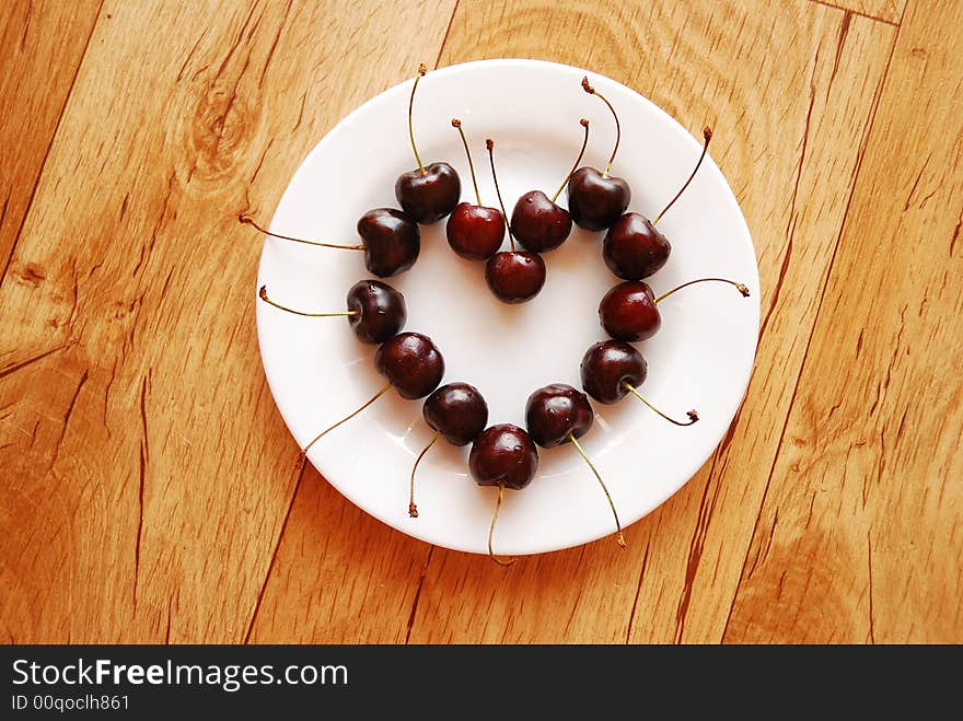 Red cherries in heart shape on the white plate, standing on the wooden table. Red cherries in heart shape on the white plate, standing on the wooden table