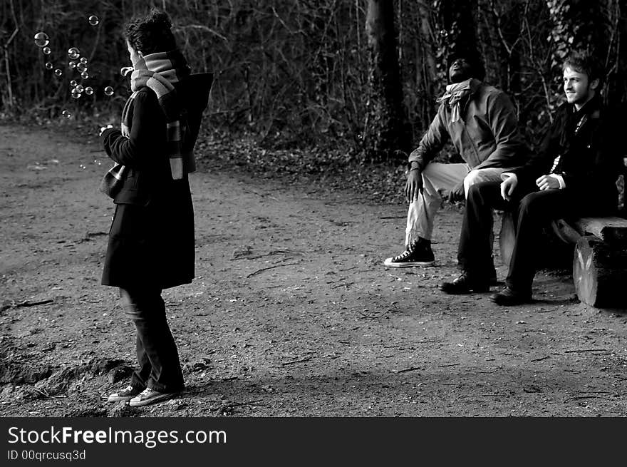 Three friends in a park: the two men are seated and the young lady is making soap bubbles. (black and white). Three friends in a park: the two men are seated and the young lady is making soap bubbles. (black and white)