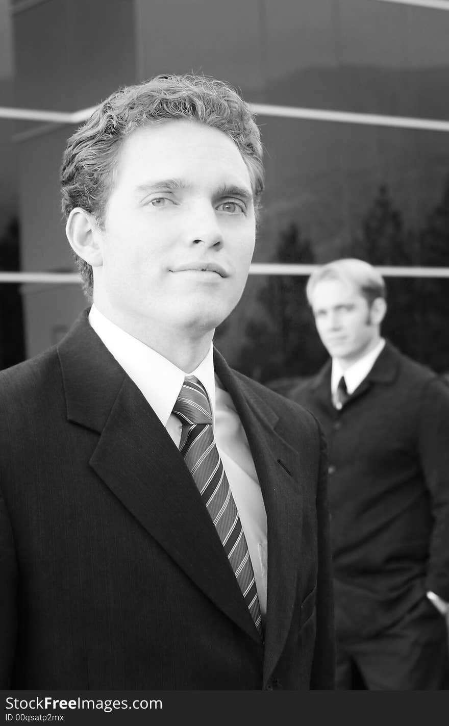 Two business men standing one in front of the other with blue office building in background. Two business men standing one in front of the other with blue office building in background