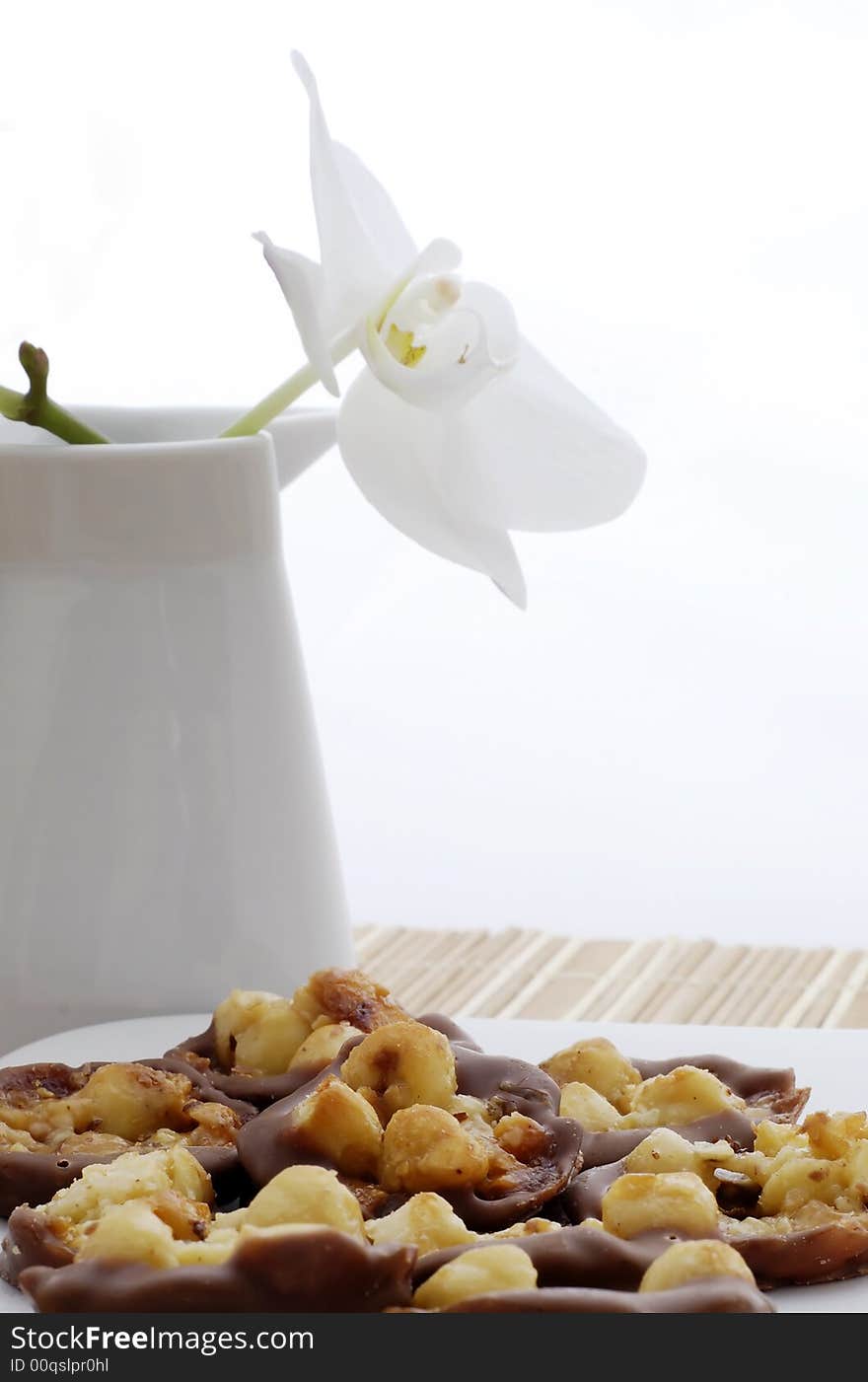 Plate of delicious nuts and chocolate with honey cakes with orchid flower in the flowerpot on background. Plate of delicious nuts and chocolate with honey cakes with orchid flower in the flowerpot on background