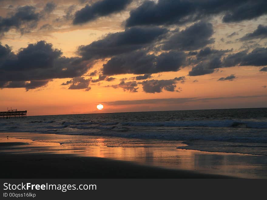 Early morning summer sunrise over ocean with sun coming up. Clouds are low and dark and sea is calm. A pier is visible in the distance at the mid-bottom left of the frame. Early morning summer sunrise over ocean with sun coming up. Clouds are low and dark and sea is calm. A pier is visible in the distance at the mid-bottom left of the frame.