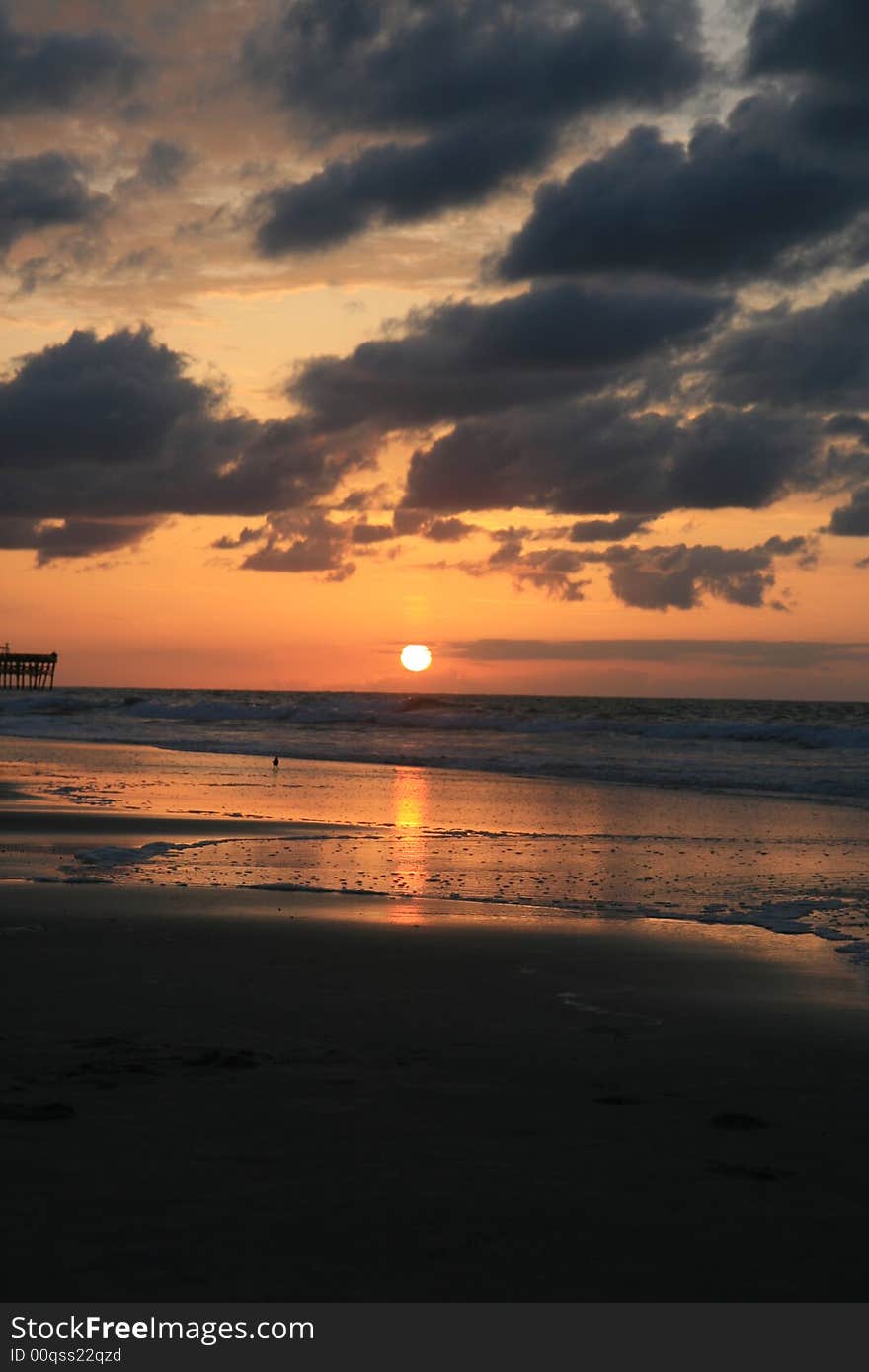 Early morning summer sunrise over ocean with sun about coming up. Clouds are low and dark and sea is calm. A pier is visible in the distance at the middle left of the frame. Early morning summer sunrise over ocean with sun about coming up. Clouds are low and dark and sea is calm. A pier is visible in the distance at the middle left of the frame.