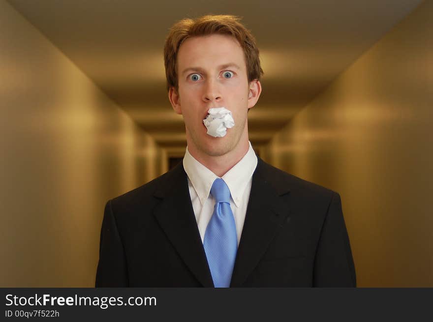 A man in a black suit stands in an empty hallway with a crumpled piece of paper in his mouth looking surprised. A man in a black suit stands in an empty hallway with a crumpled piece of paper in his mouth looking surprised