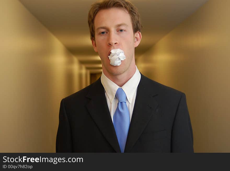 A man in a black suit stands in an empty hallway with a crumpled piece of paper in his mouth looking surprised. A man in a black suit stands in an empty hallway with a crumpled piece of paper in his mouth looking surprised