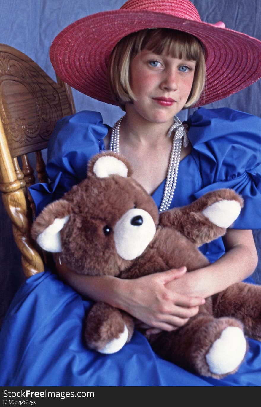 Young girl with bright blue eyes playing dress up in blue prom gown with big brimmed pink straw hat. Sitting in chair holding her brown and white teddy bear. Looking a bit forlorn. Young girl with bright blue eyes playing dress up in blue prom gown with big brimmed pink straw hat. Sitting in chair holding her brown and white teddy bear. Looking a bit forlorn.