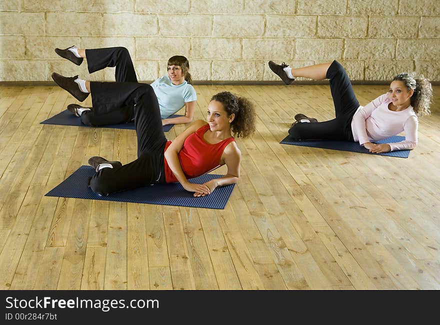 A group of women making stretching exercise on yoga mat. They're smiling and looking somewhere. Focus on the woman in red shirt. Front view. A group of women making stretching exercise on yoga mat. They're smiling and looking somewhere. Focus on the woman in red shirt. Front view.