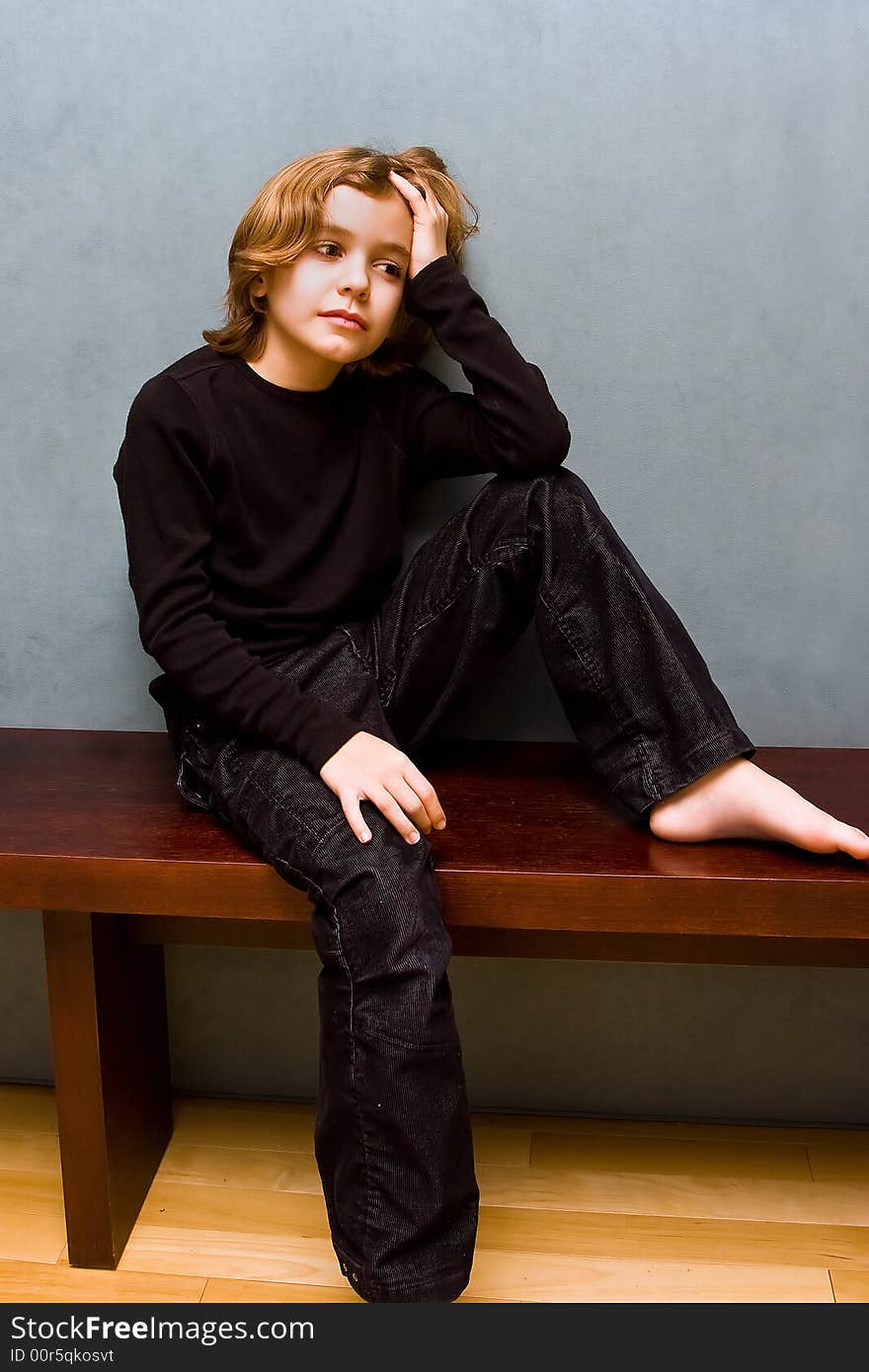 Young boy dressed in black sitting on a bench with one leg up and a hand in his hair, with a blue background. Young boy dressed in black sitting on a bench with one leg up and a hand in his hair, with a blue background