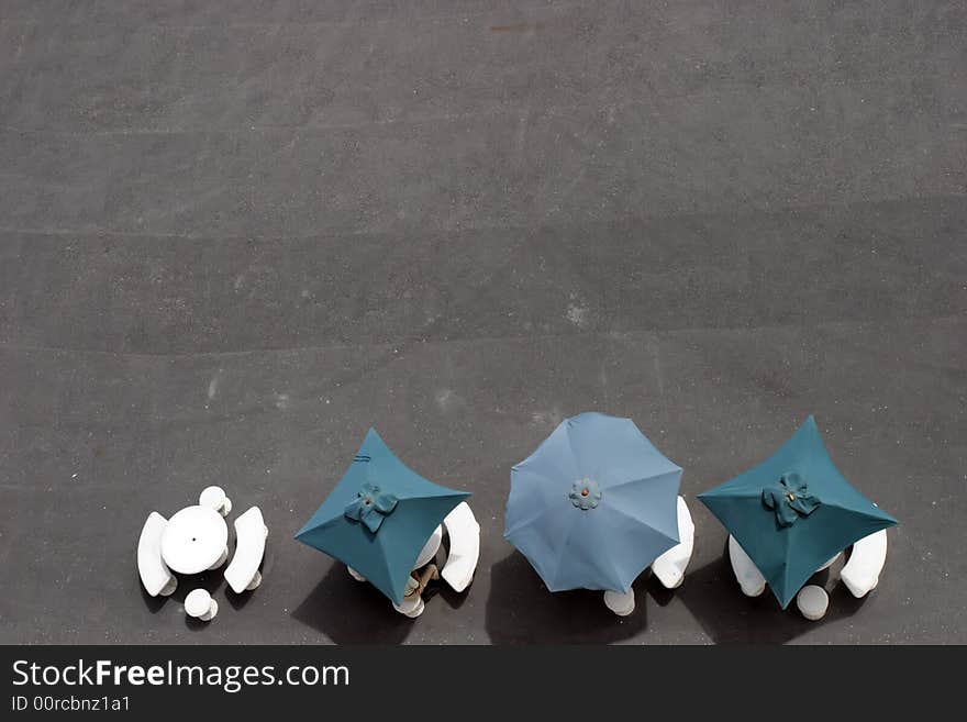 A pic of three umbrellas, four tables and a man sitting at a outdoor cafe