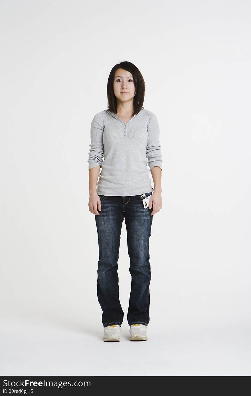 Young Asian woman with a office badge stands in a white studio wearing casual attire. She is wearing a gray shirt and denim jeans with a blank look on her face. Young Asian woman with a office badge stands in a white studio wearing casual attire. She is wearing a gray shirt and denim jeans with a blank look on her face.