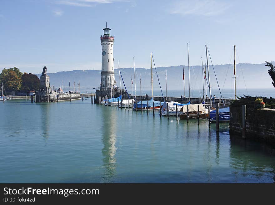 Lindau harbour looking at the harbour entrance guarded by a lighthouse and statue of a lion. Sailing boats are moored along the breakwater and the lighthouse is reflected in the calm water. Lake Constance is covered in early morning mist. Lindau harbour looking at the harbour entrance guarded by a lighthouse and statue of a lion. Sailing boats are moored along the breakwater and the lighthouse is reflected in the calm water. Lake Constance is covered in early morning mist.