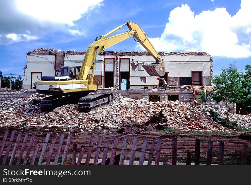 Yellow bulldozer turning old building into rubble beneath a brilliant summer sky. Huge debris pile and construction fence in foreground. Yellow bulldozer turning old building into rubble beneath a brilliant summer sky. Huge debris pile and construction fence in foreground.