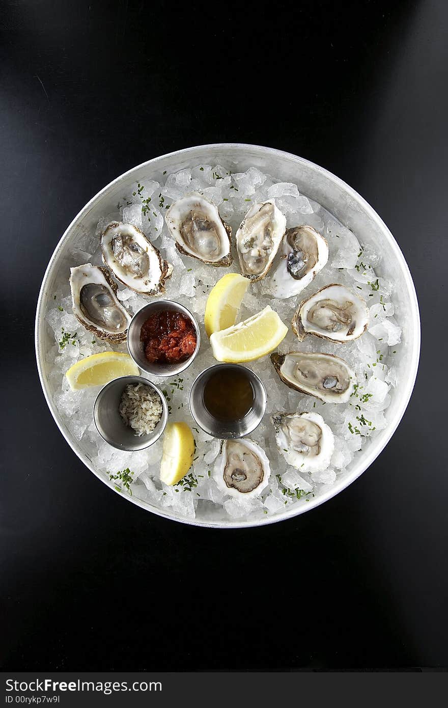 A seafood appetizer of clams on a black table shot from above. The clams are in a tray of ice with lemons and sauce.