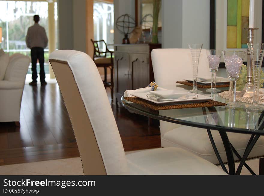 In the foreground, a white chair is pushed into a glass dining room table. The table is set with white and crystal dishes. A man stands in the background.