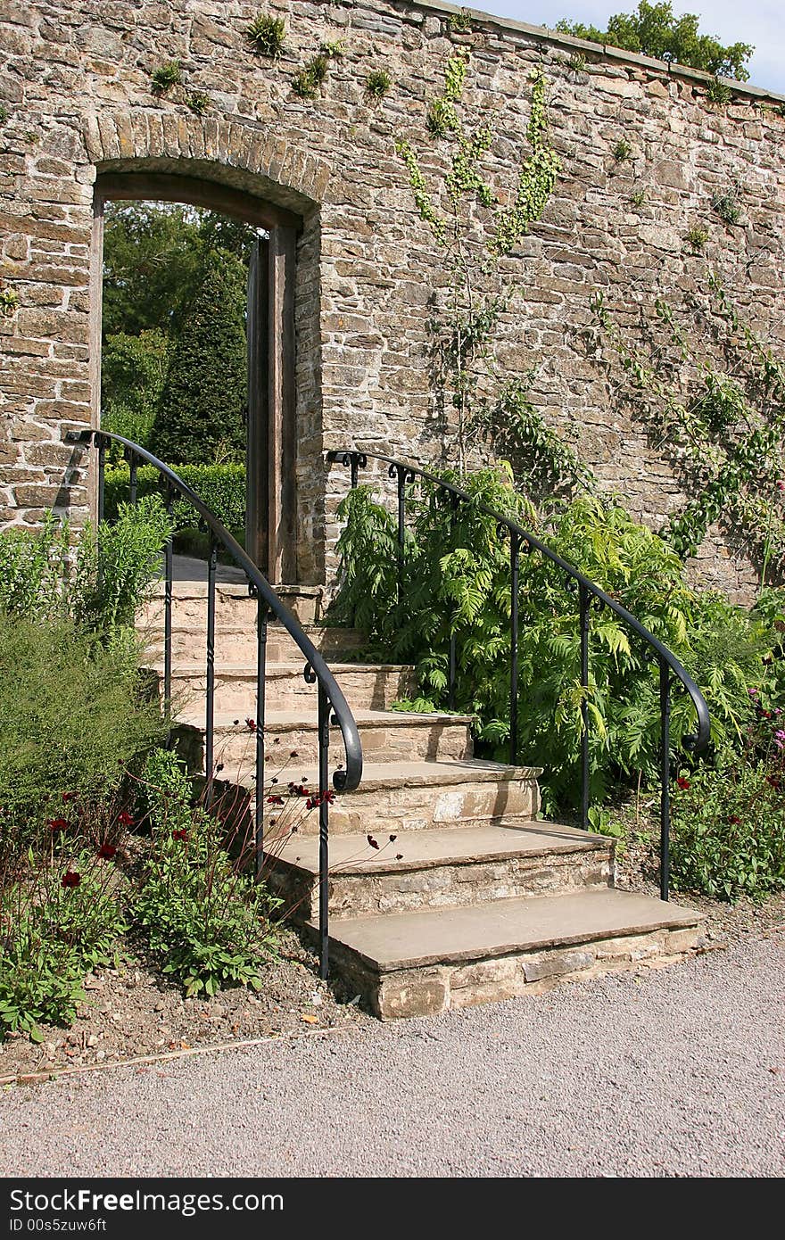 Ancient garden stairway with metal handrails, set within an old stone wall and leading to a garden beyond. Flowers and shrubs either side of the steps.