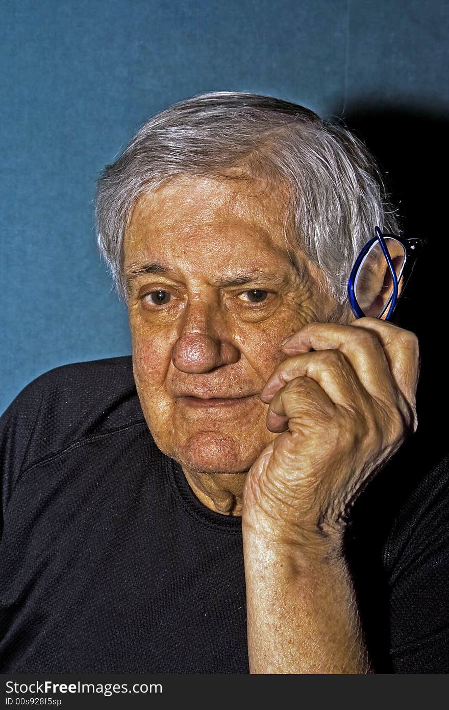 Dramatic side portrait of a senior man with a pair of glasses in his hand next to his face wearing a black shirt isolated on gray/blue. Dramatic side portrait of a senior man with a pair of glasses in his hand next to his face wearing a black shirt isolated on gray/blue