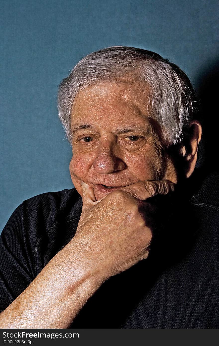 Dramatic portrait of a senior man with his hand on the side of his face wearing a black shirt isolated on gray/blue