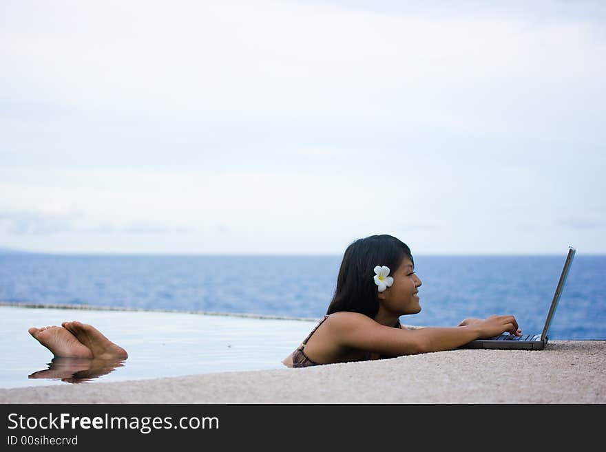 Attractive young Asian business woman working remotely on a laptop computer beside an infinity pool at a private tropical resort paradise vacation. Room for text. Attractive young Asian business woman working remotely on a laptop computer beside an infinity pool at a private tropical resort paradise vacation. Room for text.