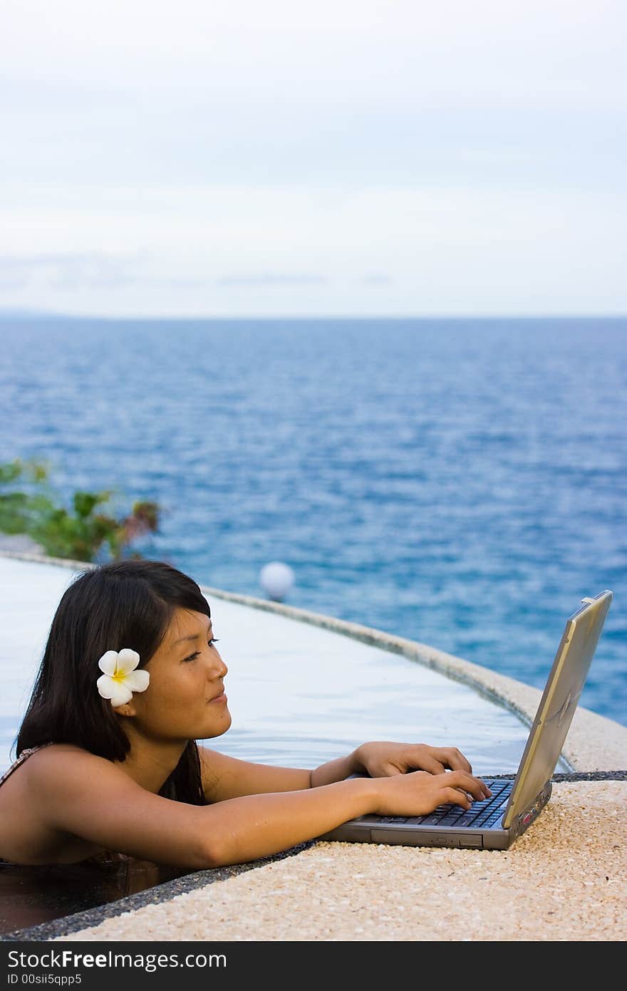 Attractive young Asian business woman working remotely on a laptop computer beside an infinity pool at a private tropical resort paradise vacation. Room for text. Attractive young Asian business woman working remotely on a laptop computer beside an infinity pool at a private tropical resort paradise vacation. Room for text.