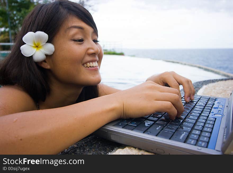 Attractive young Asian business woman working remotely on a laptop computer beside an infinity pool at a private tropical resort paradise vacation. Room for text. Attractive young Asian business woman working remotely on a laptop computer beside an infinity pool at a private tropical resort paradise vacation. Room for text.