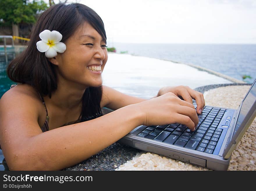 Attractive young Asian business woman working remotely on a laptop computer beside an infinity pool at a private tropical resort paradise vacation. Room for text. Attractive young Asian business woman working remotely on a laptop computer beside an infinity pool at a private tropical resort paradise vacation. Room for text.