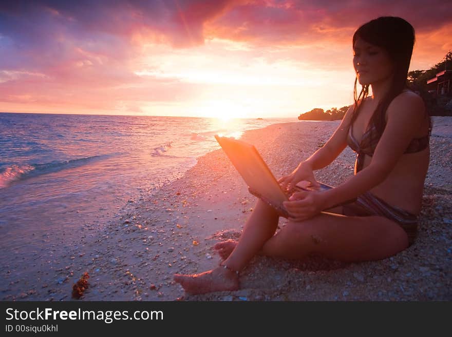 Attractive young Asian business woman working remotely on a laptop computer on a private white sand beach beside the ocean at tropical resort paradise vacation with beautiful red sunset background. Room for text. Attractive young Asian business woman working remotely on a laptop computer on a private white sand beach beside the ocean at tropical resort paradise vacation with beautiful red sunset background. Room for text.