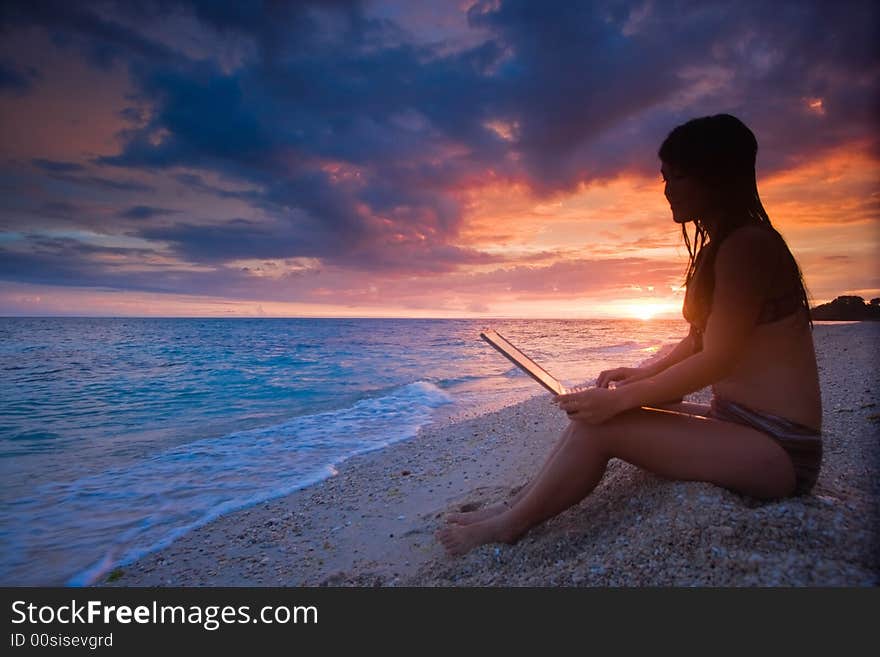 Attractive young Asian business woman working remotely on a laptop computer on a private white sand beach beside the ocean at tropical resort paradise vacation with beautiful red sunset background. Room for text. Attractive young Asian business woman working remotely on a laptop computer on a private white sand beach beside the ocean at tropical resort paradise vacation with beautiful red sunset background. Room for text.