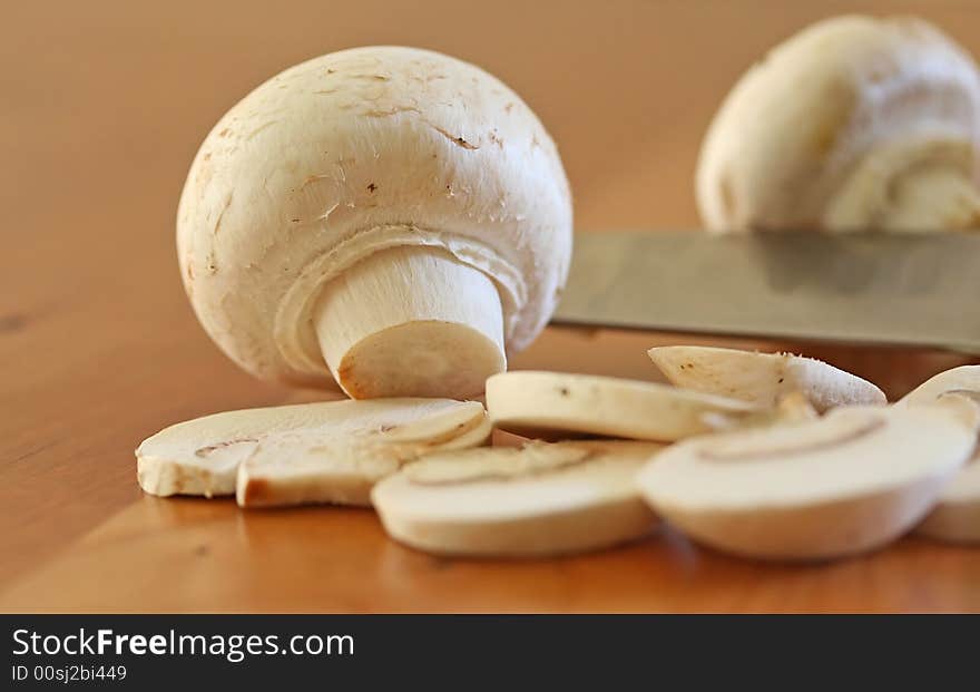 Mushrooms being sliced on a wood cutting board with stainless steel knife edge in frame. Mushrooms being sliced on a wood cutting board with stainless steel knife edge in frame.