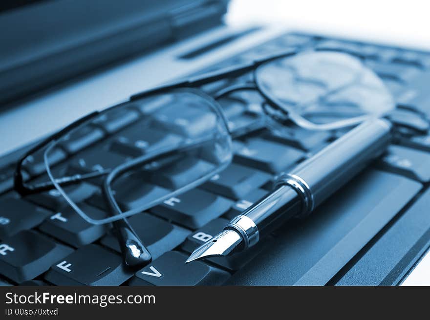 A pair of glasses and a fountain pen on a laptop on blue. Symbol of business working in office. Soft focus. A pair of glasses and a fountain pen on a laptop on blue. Symbol of business working in office. Soft focus.