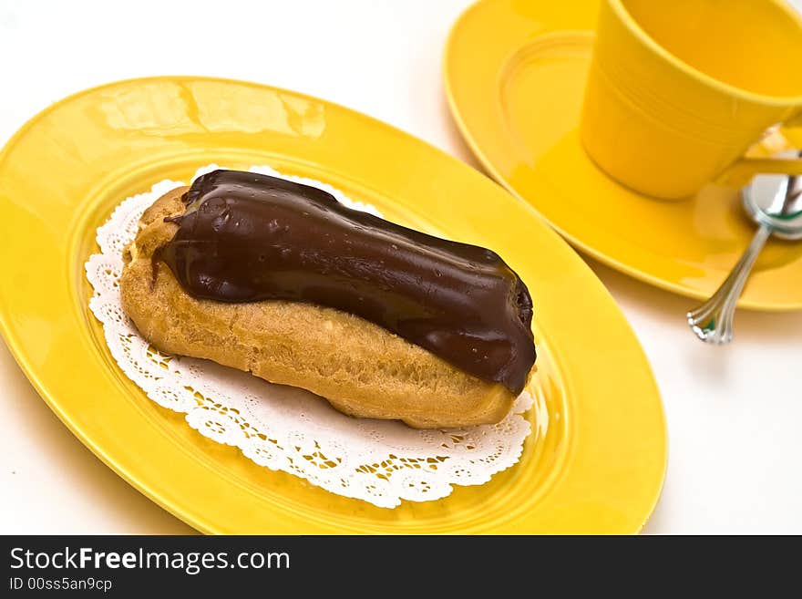 A chocolate eclair on yellow plate with coffee cup and saucer.