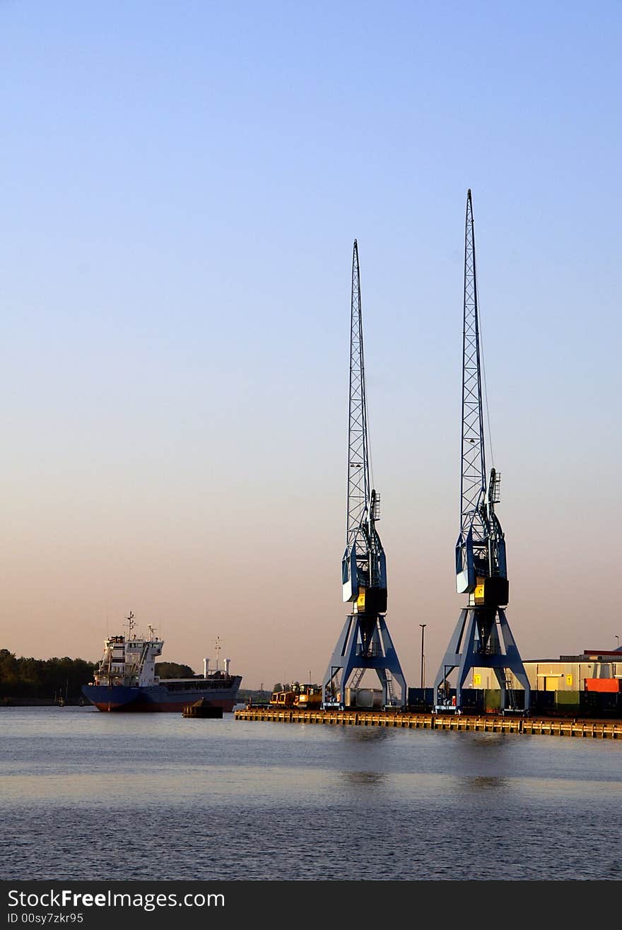 Two similar blue harbor cranes waiting for the next shipload. A ship is passing by. There is cargo on the shore.