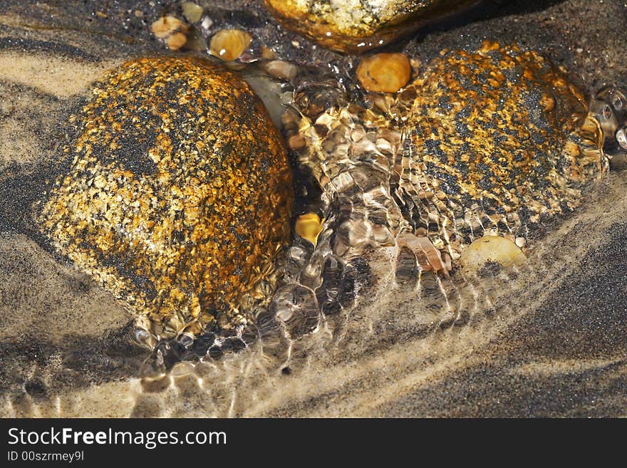 Rocks and pebbles glisten in a rivulet of water. The warm colors of the rocks and pebbles are contrasted against the cooler tones of the striated sandy bed. Rocks and pebbles glisten in a rivulet of water. The warm colors of the rocks and pebbles are contrasted against the cooler tones of the striated sandy bed.