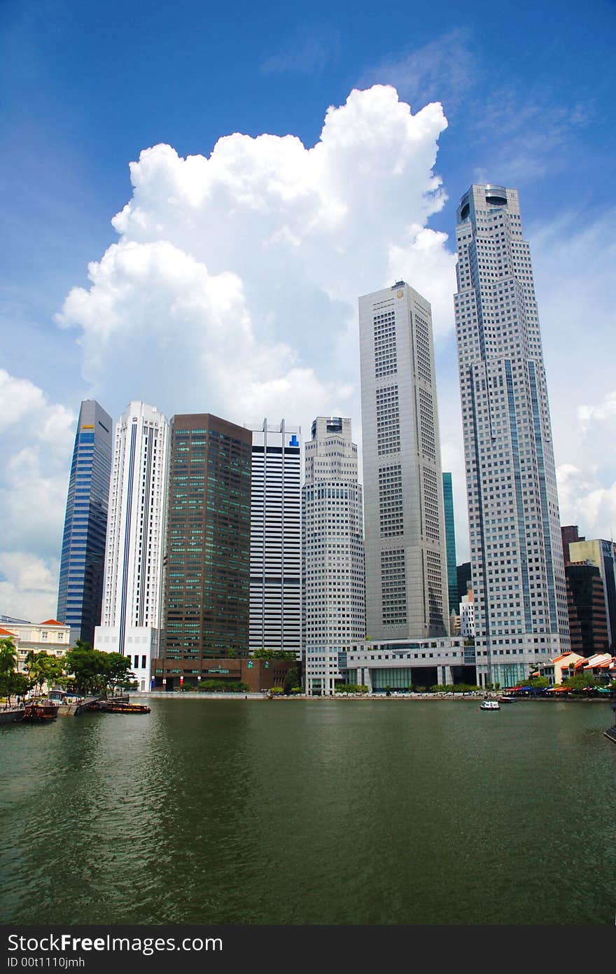 Towering skyscrapers loom over a reflective river and surrounded by huge clouds.