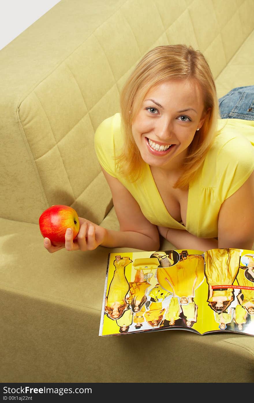 The girl lays on a sofa, reads the cooking magazine and eats an apple. The girl lays on a sofa, reads the cooking magazine and eats an apple