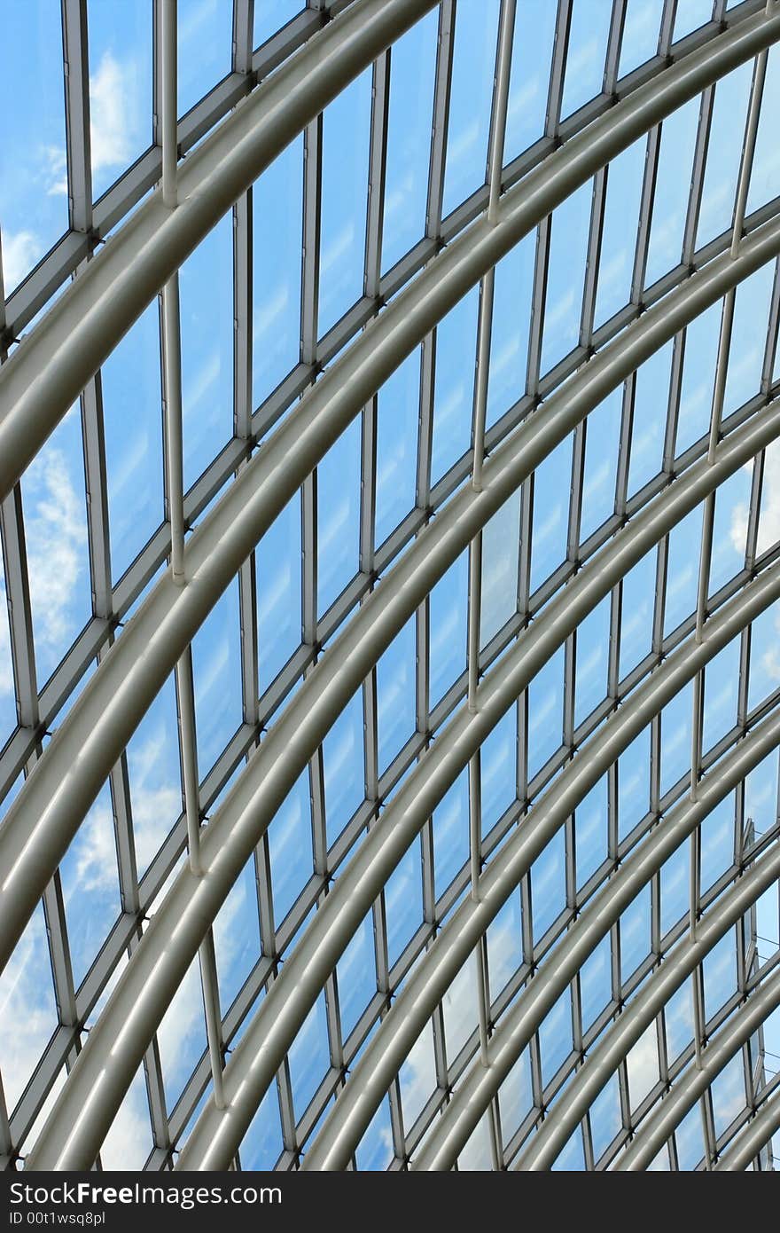 Silver metal curved roof joists in a conservatory with glass panes in between and a blue sky and clouds beyond. Silver metal curved roof joists in a conservatory with glass panes in between and a blue sky and clouds beyond.