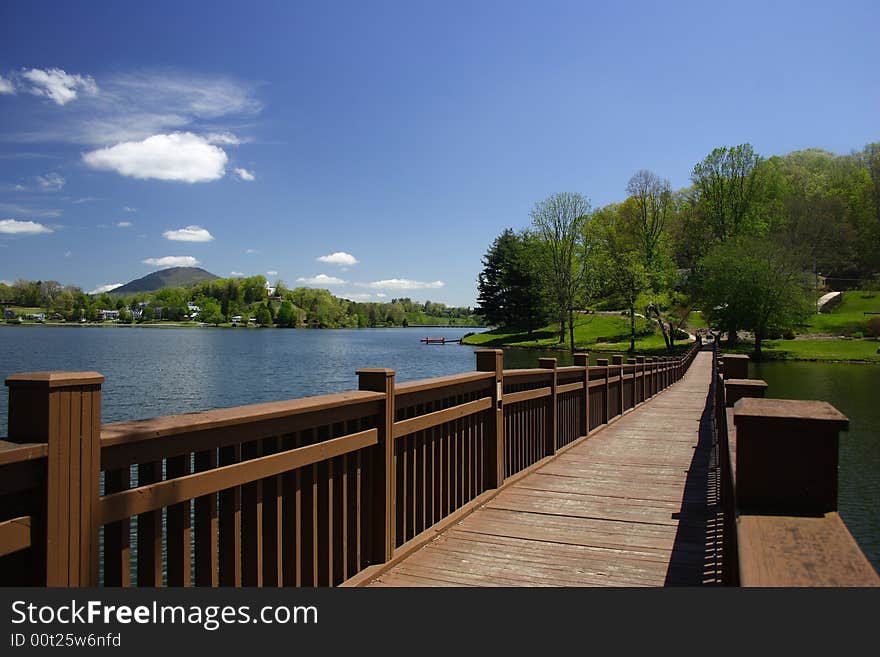 A footbridge leading across a small lake with trees and a blue sky in the background