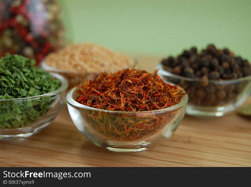 Glass containers with saffron, mustard seed, black pepper and chive and a bottle of mixed pepper on a wooden cutting desk with a green background. Glass containers with saffron, mustard seed, black pepper and chive and a bottle of mixed pepper on a wooden cutting desk with a green background