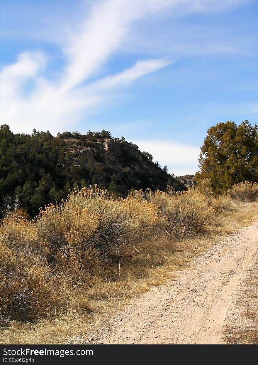 A dirt road leading up a hill with dried bushes, a hill and blue sky in the background. A dirt road leading up a hill with dried bushes, a hill and blue sky in the background.