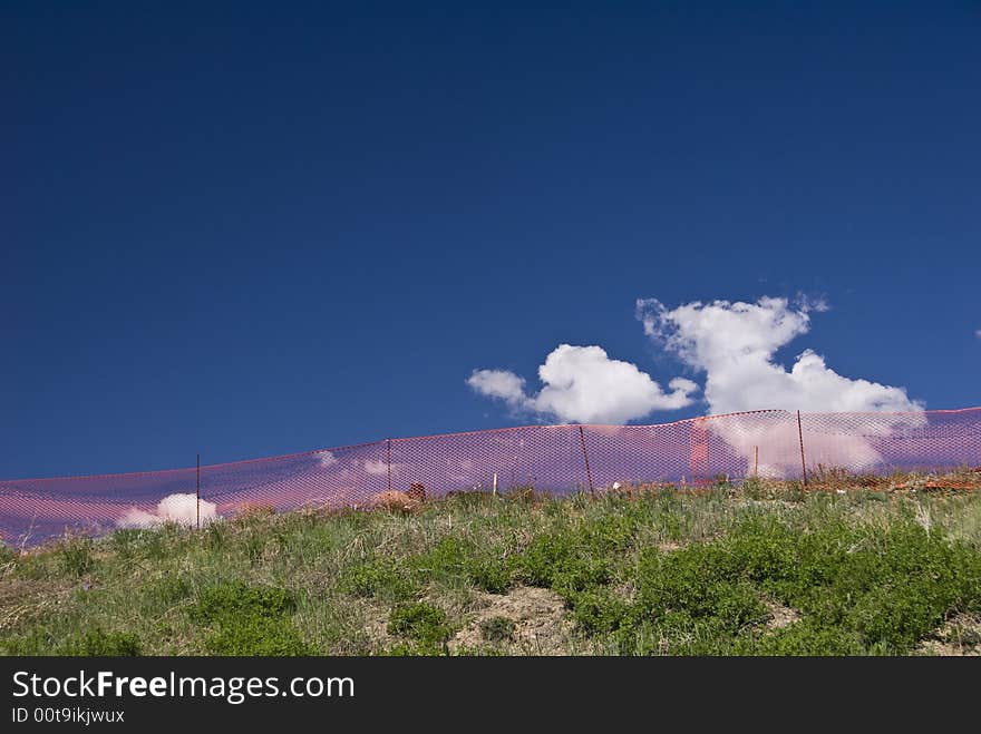 Orange construction fence on grassy hill with blue sky and white puffy cloud