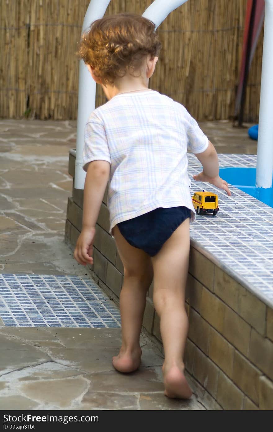 A curly little boy plays with the toy car of white and yellow color at a pool. A curly little boy plays with the toy car of white and yellow color at a pool