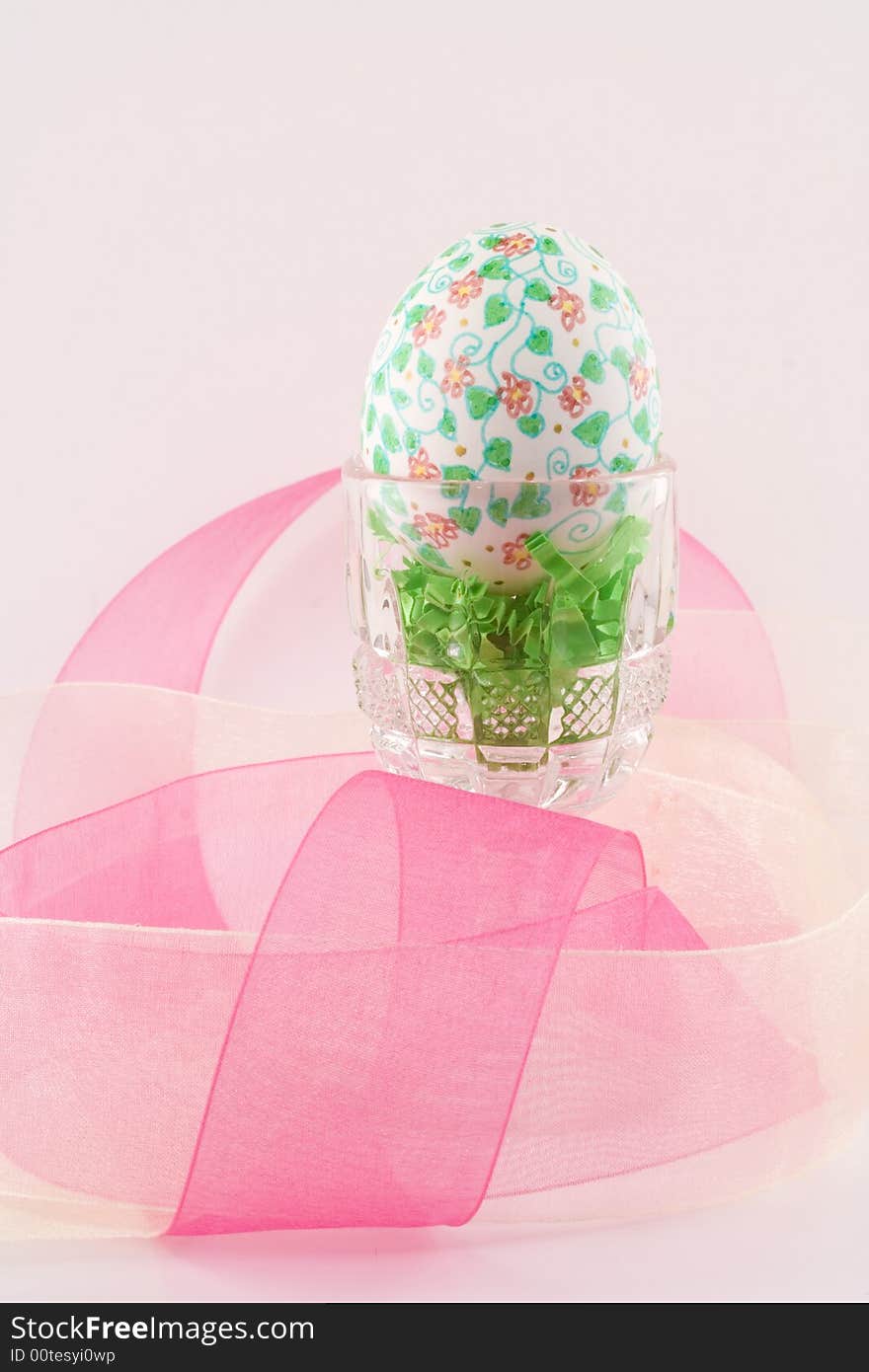 A single hand decorated Easter egg sits in a crystal cup surrounded by pink and yellow organza ribbon over a pale pink background.