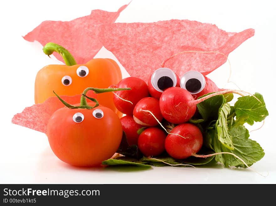 A bunch of radishes, a tomato and an orange bell pepper with googly eyes and red capes sit on a white background. A bunch of radishes, a tomato and an orange bell pepper with googly eyes and red capes sit on a white background.